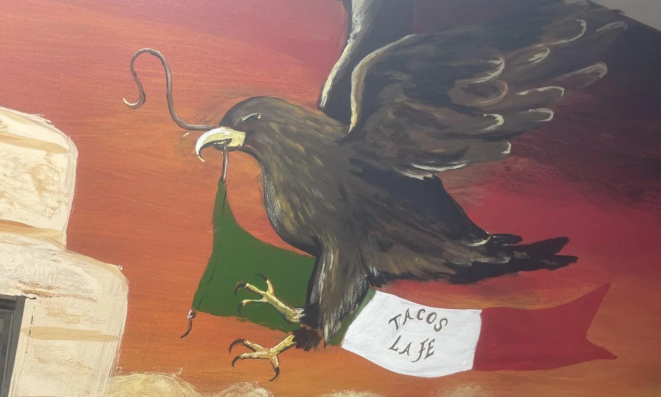 A painted mural of a Mexican eagle holding a snake over the flag, with a banner reading Tacos La Fe