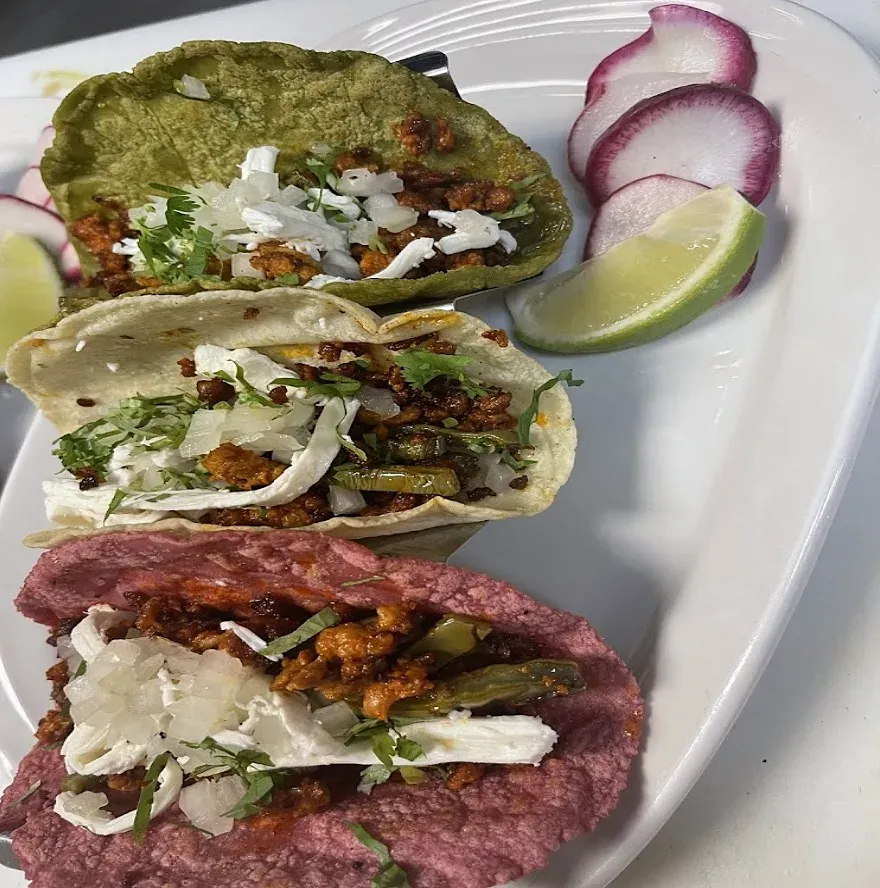 Three tacos on green, white and red tortillas with onion and cilantro, beside radish slices and a lime