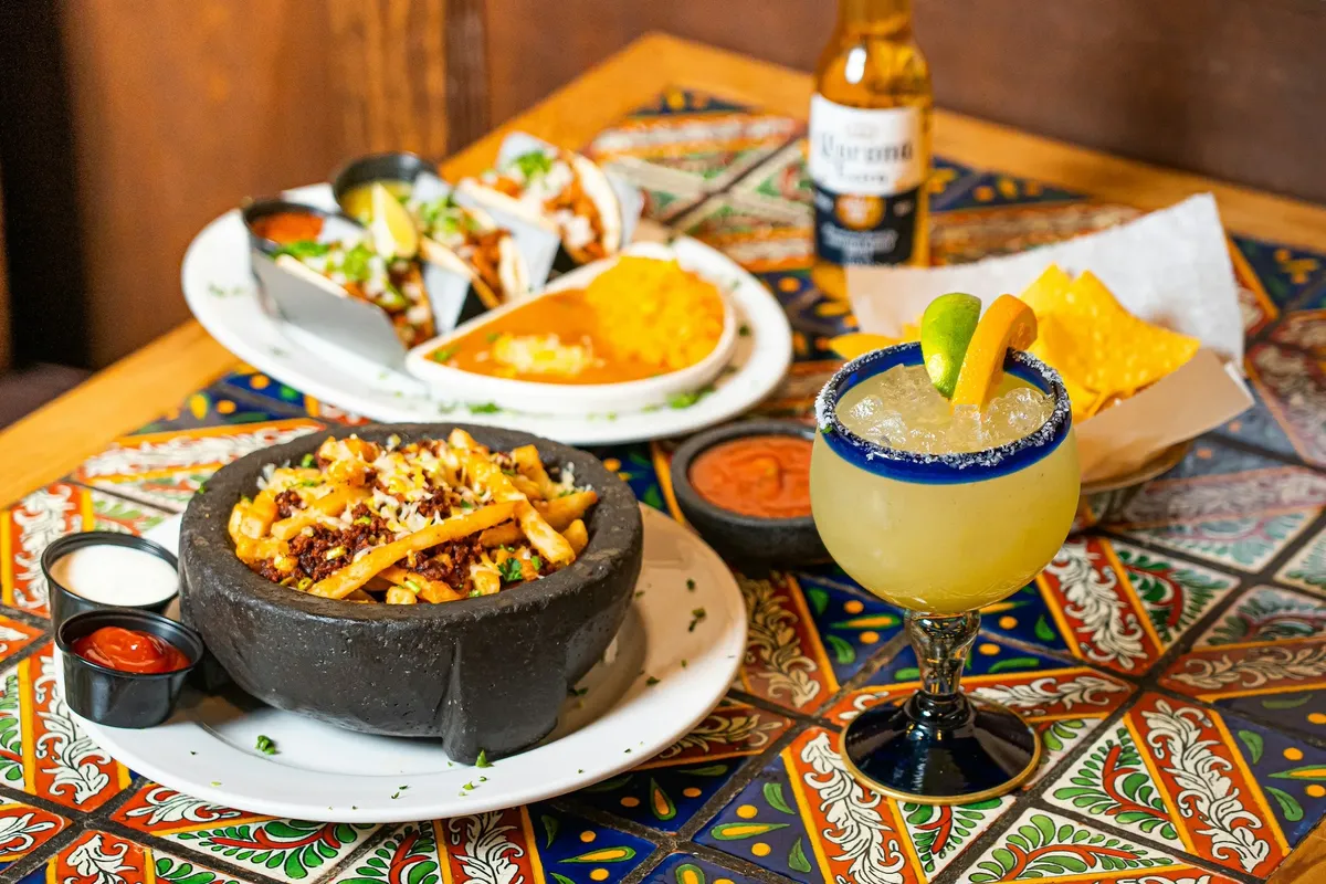 A molcajete of fries topped with meat and cheese, tacos and a salt-rimmed margarita on a talavera-tiled table