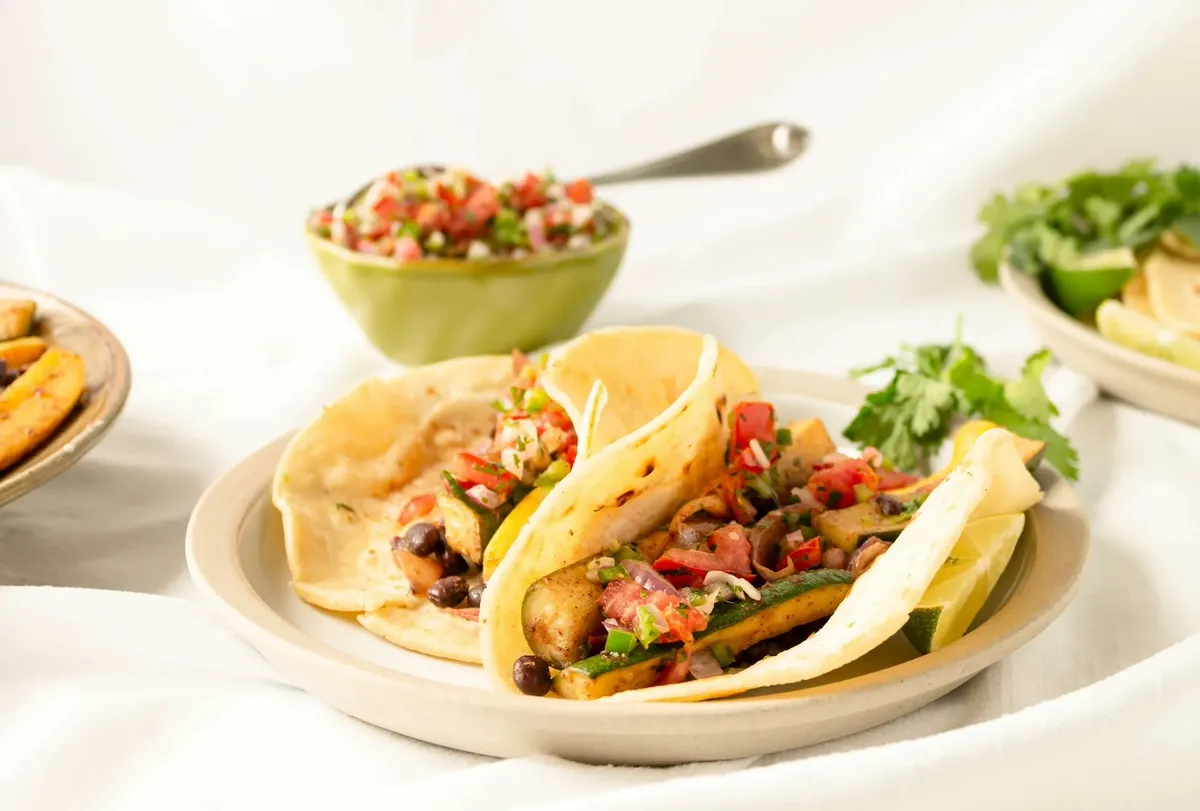 Two tacos with pico de gallo beside a bowl of salsa on a white table