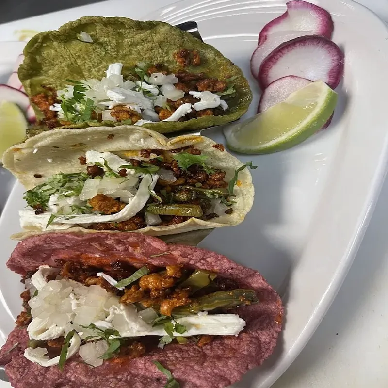 Three tacos on green, white and red tortillas with onion and cilantro, beside radish slices and a lime