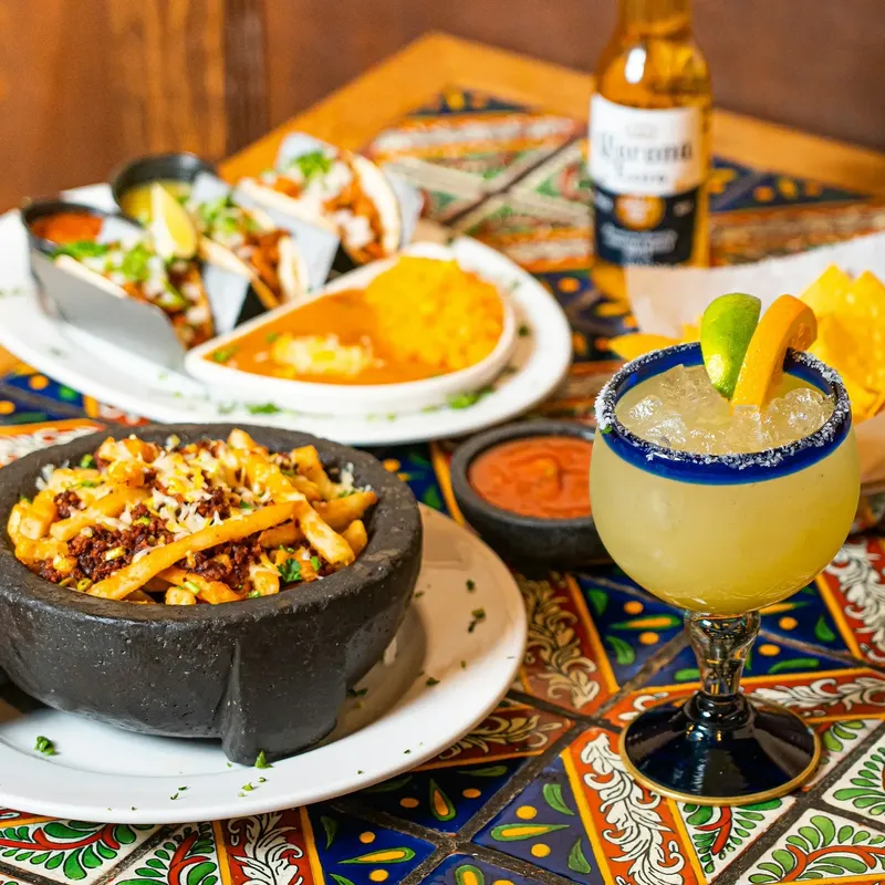A molcajete of fries topped with meat and cheese, tacos and a salt-rimmed margarita on a talavera-tiled table