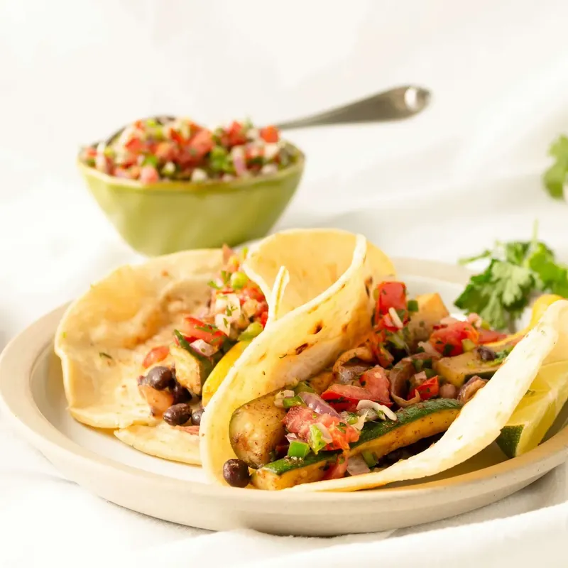 Two tacos with pico de gallo beside a bowl of salsa on a white table